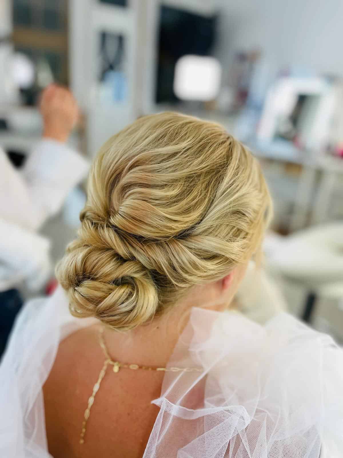 Eine Frau mit blonden Haaren, die zu einem eleganten, gedrehten, tief sitzenden Dutt frisiert sind, trägt eine zarte goldene Halskette und ein weißes Tüllkleid und sitzt in einem hellen, modernen Salon – einem einladenden Raum, der an die gehobene Atmosphäre während einer Kosmetikerinnenausbildung an der renommierten Kosmetikschule Einbeck erinnert.