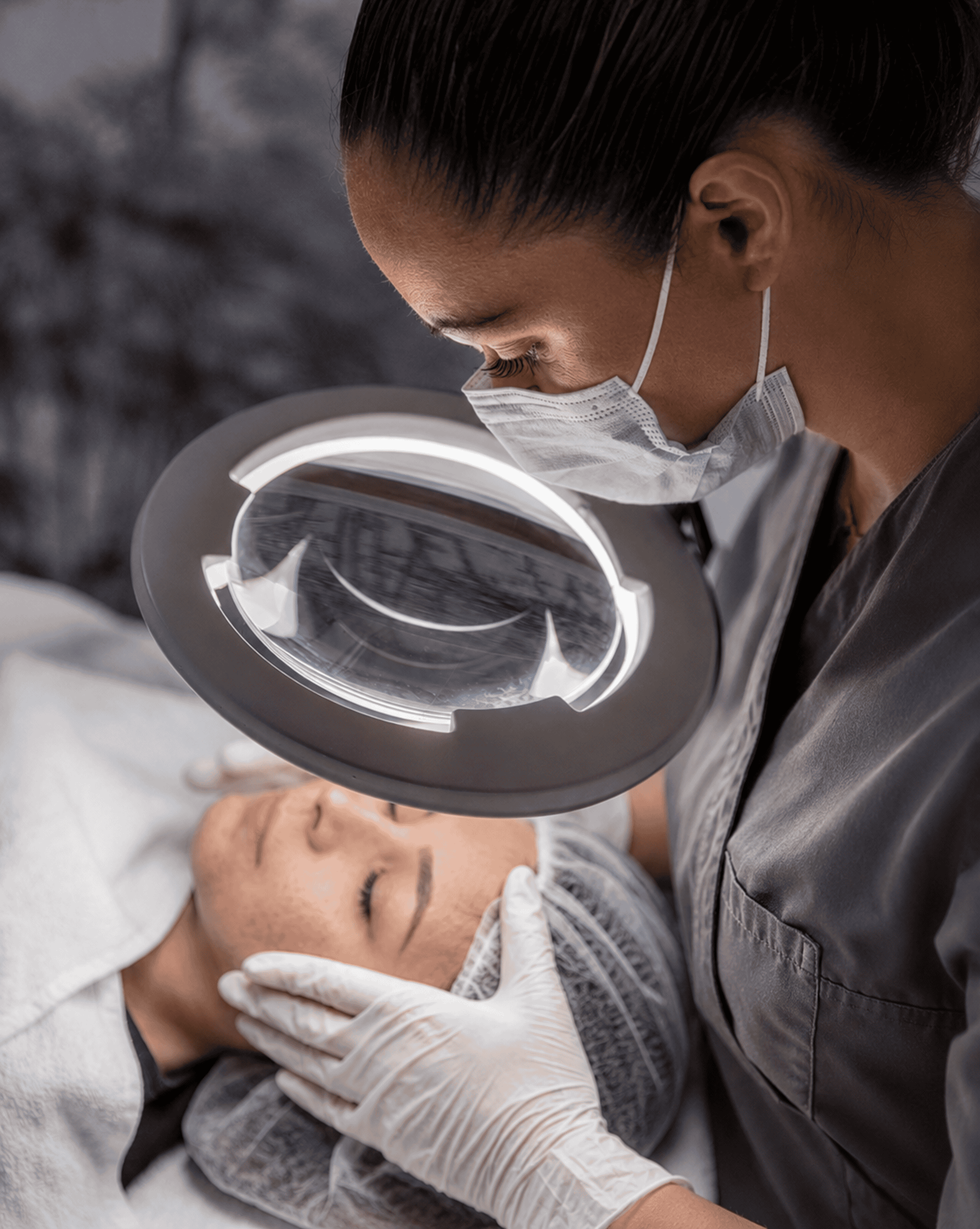 Eine Hautpflegeexpertin, die eine Kosmetikerinnenausbildung absolviert hat, untersucht mit Handschuhen und Maske das Gesicht einer Patientin unter einer Lupenlampe. Die Patientin liegt entspannt mit geschlossenen Augen da, bedeckt mit einem Haarnetz und einem Handtuch – eine eindringliche Szene aus der renommierten Kosmetikschule Einbeck oder einer professionellen Kosmetikpraxis in Einbeck.