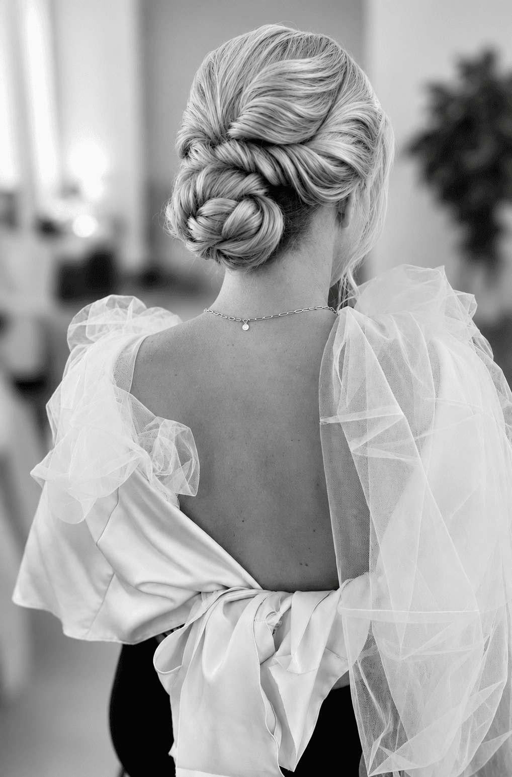 Eine Frau mit einer eleganten Hochsteckfrisur und einer zarten Halskette ist von hinten zu sehen; sie trägt ein Kleid mit Tüllverzierungen und einem offenen Rücken. Das Bild, das an die zurückhaltende Eleganz erinnert, die an der Kosmetikschule Einbeck gepflegt wird, ist in Schwarz-Weiß gehalten.
