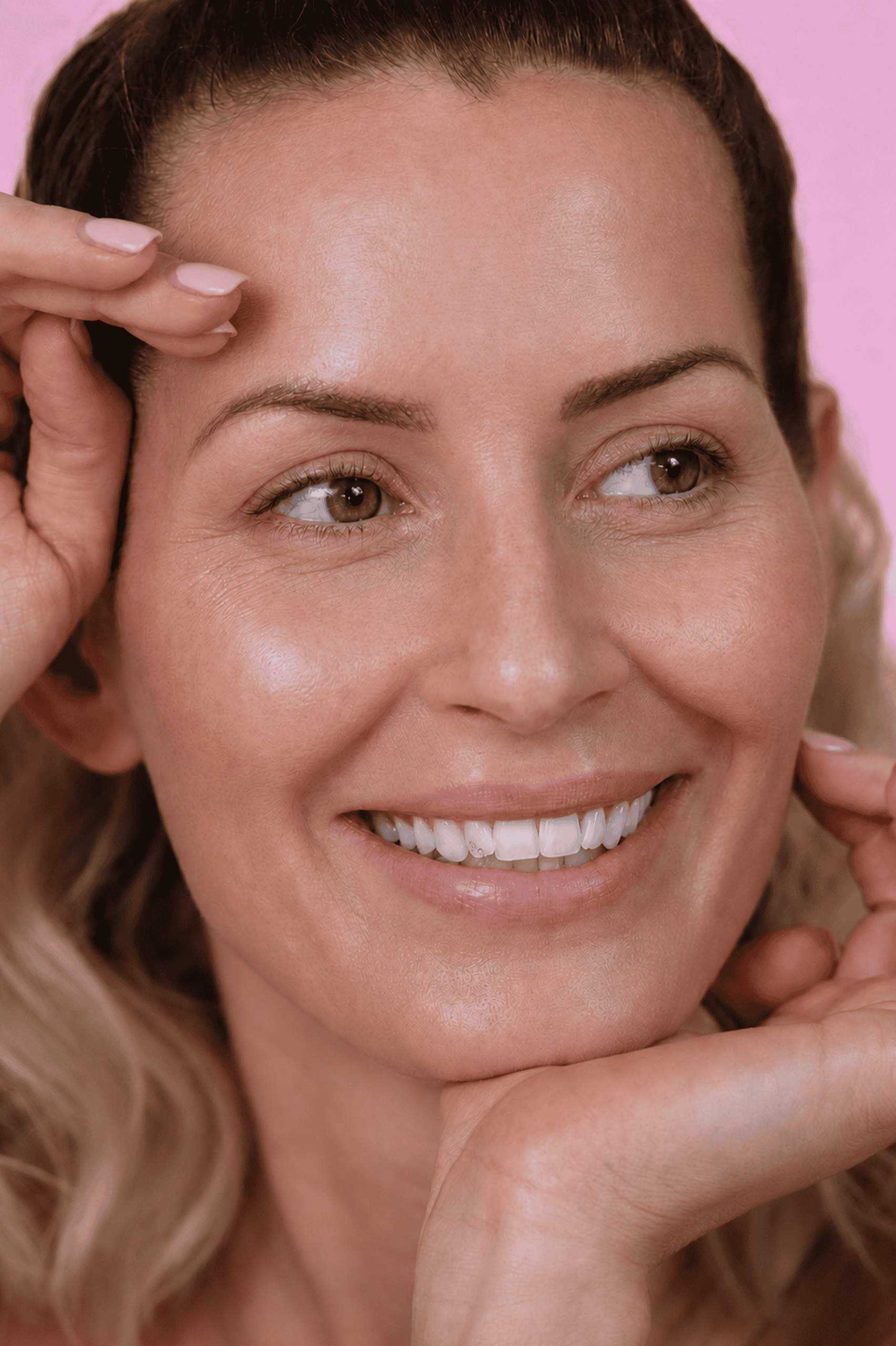 Eine Frau mit welligem blondem Haar lächelt und berührt ihr Gesicht mit beiden Händen. Sie hat reine Haut, trägt dezentes Make-up und steht vor einem rosa Hintergrund – ein frischer, professioneller Look, wie man ihn oft an der Kosmetikschule Einbeck sieht.