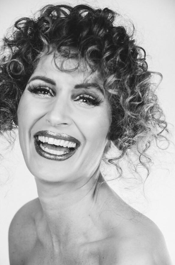 Schwarz-Weiß-Foto einer Frau mit lockigem Haar, die breit lächelt und dabei ihre Zähne zeigt. Sie trägt auffälliges Augen-Make-up und glänzenden Lippenstift, was den künstlerischen Stil widerspiegelt, wie er oft in der Kosmetikpraxis Einbeck zu sehen ist. Der Hintergrund ist schlicht und hell.
