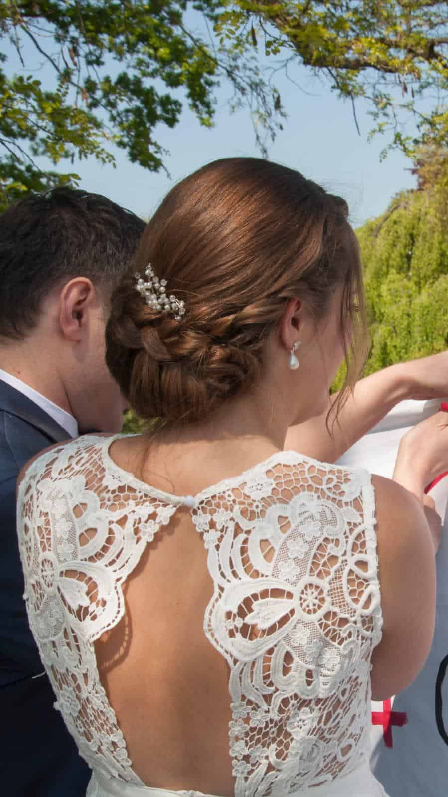 Eine Frau in einem weißen Spitzenkleid mit einer aufwendig gestylten Hochsteckfrisur – vielleicht das Ergebnis einer professionellen Kosmetikausbildung – steht im Freien neben einem Mann im Anzug; beide haben dem Betrachter den Rücken zugewandt und blicken auf etwas, das außerhalb des Bildausschnitts liegt. Im Hintergrund sind Bäume und Grünflächen zu sehen.