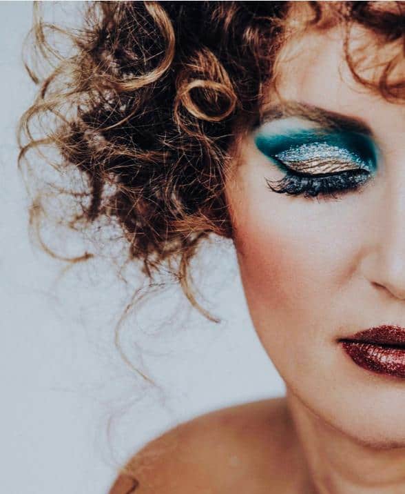 Nahaufnahme einer Person mit lockigem Haar, auffälligem Augen-Make-up in Blaugrün und Gold, langen Wimpern und dunkelrotem Lippenstift, gezeigt von den Schultern aufwärts vor einem hellen Hintergrund. Nur die Hälfte ihres Gesichts ist zu sehen – ein Beispiel für die künstlerische Kunstfertigkeit, die in einer professionellen Kosmetikausbildung vermittelt wird, wie sie beispielsweise an der Kosmetikschule Einbeck angeboten wird.