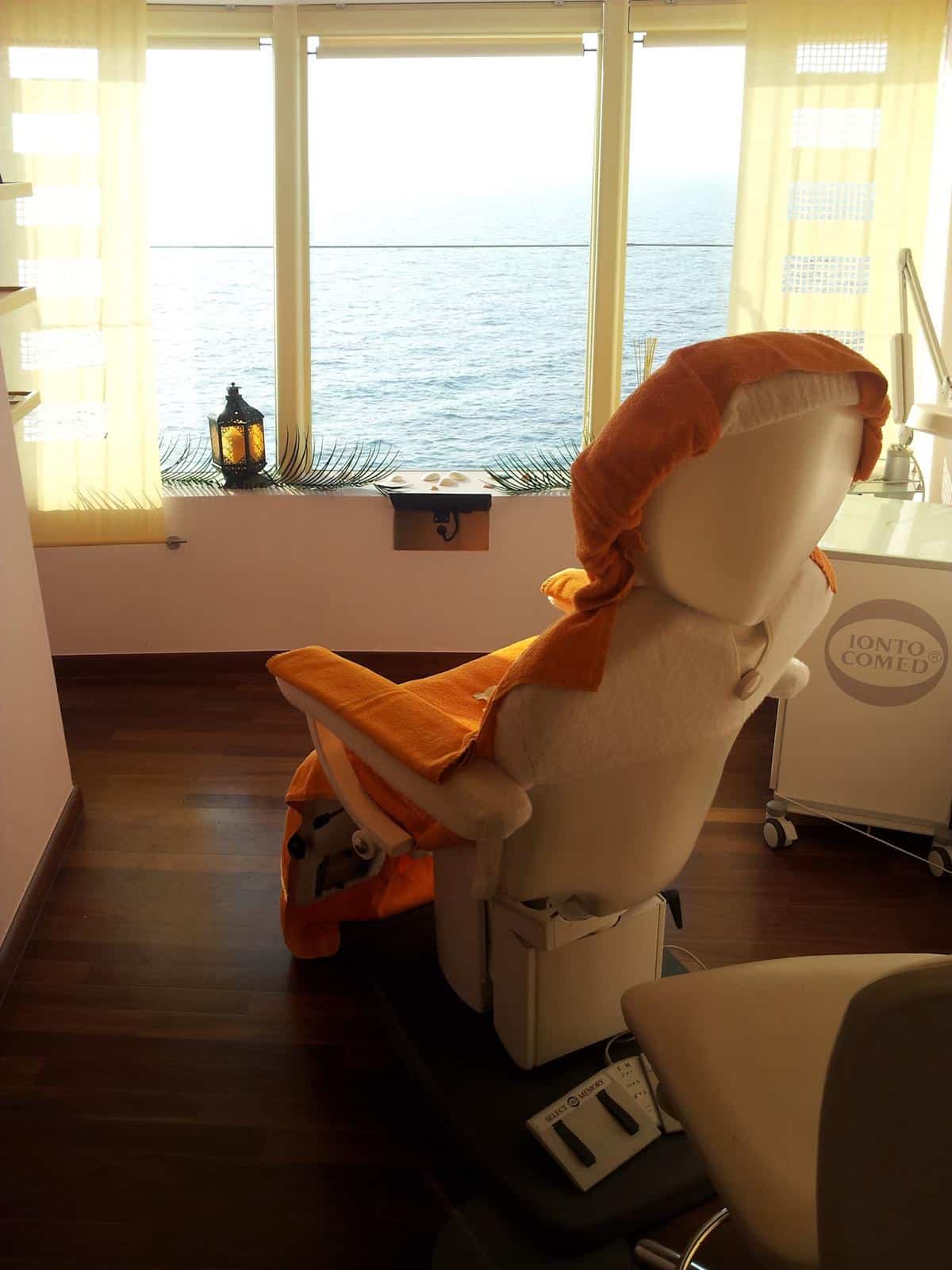 Ein mit orangefarbenen und weißen Handtüchern bezogener Wellness-Behandlungsstuhl steht vor großen Fenstern mit Blick auf das Meer, durch die das Sonnenlicht hereinströmt, und auf der Fensterbank steht eine Laterne – ein einladender Raum, der an die Schulungsräume einer Kosmetik-Privatschule oder an die ruhigen Sitzungen während Ihrer Ausbildung zur Kosmetikerin erinnert.