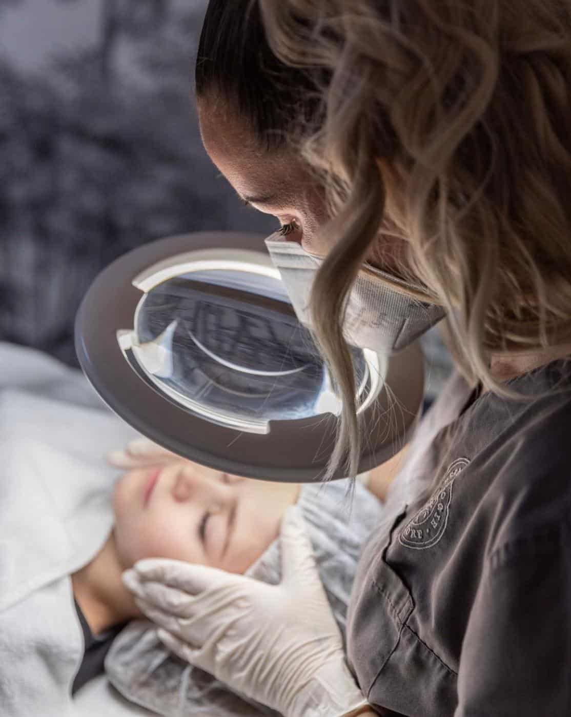 Eine Person, die Handschuhe und eine Maske trägt, untersucht das Gesicht einer anderen Person unter einer Lupenlampe, was auf eine Hautpflege- oder Gesichtsbehandlung in einer Klinik oder einem Spa hindeutet – vielleicht im Rahmen eines praktischen Trainings während einer Kosmetikerin-Ausbildung oder an der Kosmetikschule Einbeck.