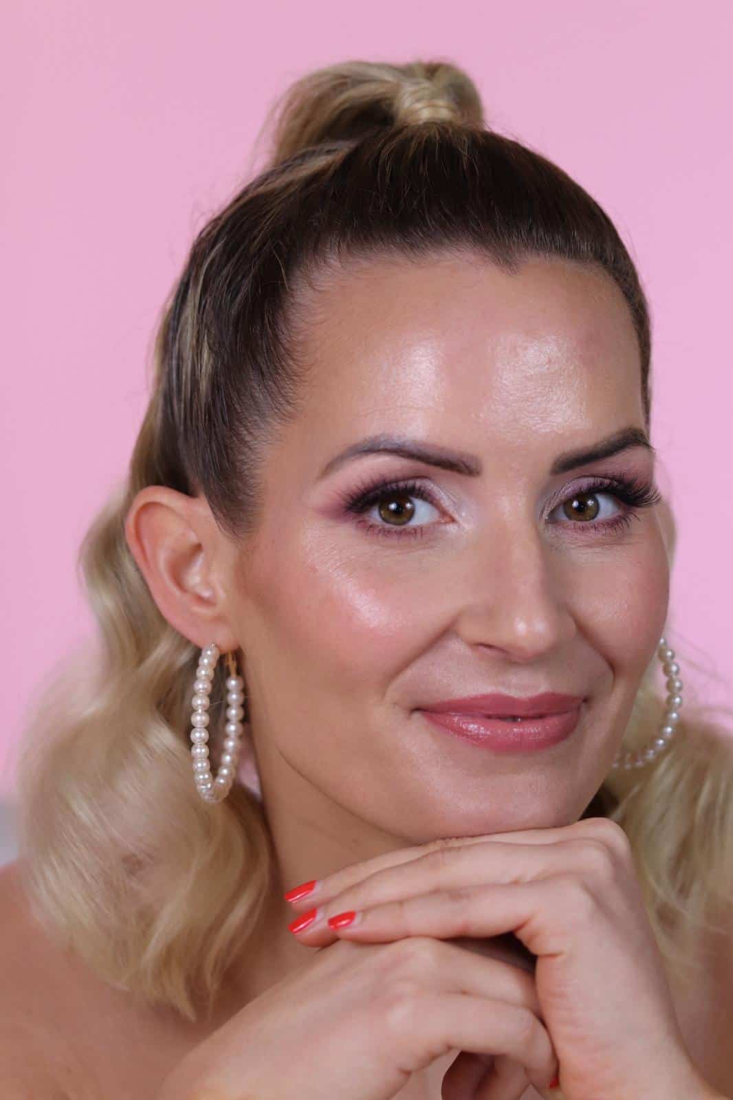 Eine Frau mit welligem blondem Haar, Perlen-Creolen und rosa Make-up lächelt sanft vor einem rosa Hintergrund, während sie ihr Kinn auf ihre Hände mit rot lackierten Nägeln stützt – ein Bild, das die Eleganz und Professionalität, die durch die Kosmetikerinnenausbildung an der Kosmetikschule Einbeck vermittelt werden, wunderbar einfängt.