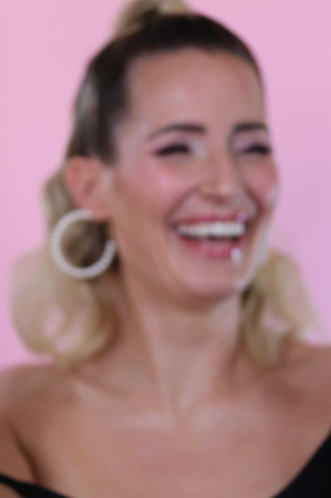 Ein unscharfes Bild einer lächelnden Frau mit hellem Haar, die große Creolen und ein schulterfreies Oberteil trägt und vor einem rosa Hintergrund zu sehen ist, vermittelt die lebendige und einladende Atmosphäre der Kosmetikschule Einbeck.