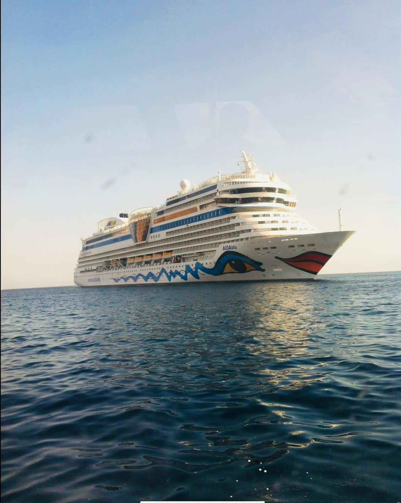 Ein großes weißes Kreuzfahrtschiff mit blauen und gelben Wellenmustern und einem rot umrandeten Lächeln auf dem Bug schwimmt unter klarem Himmel auf ruhigem, offenem Wasser und erinnert an die leuchtenden Akzente in der Kosmetikpraxis Einbeck.