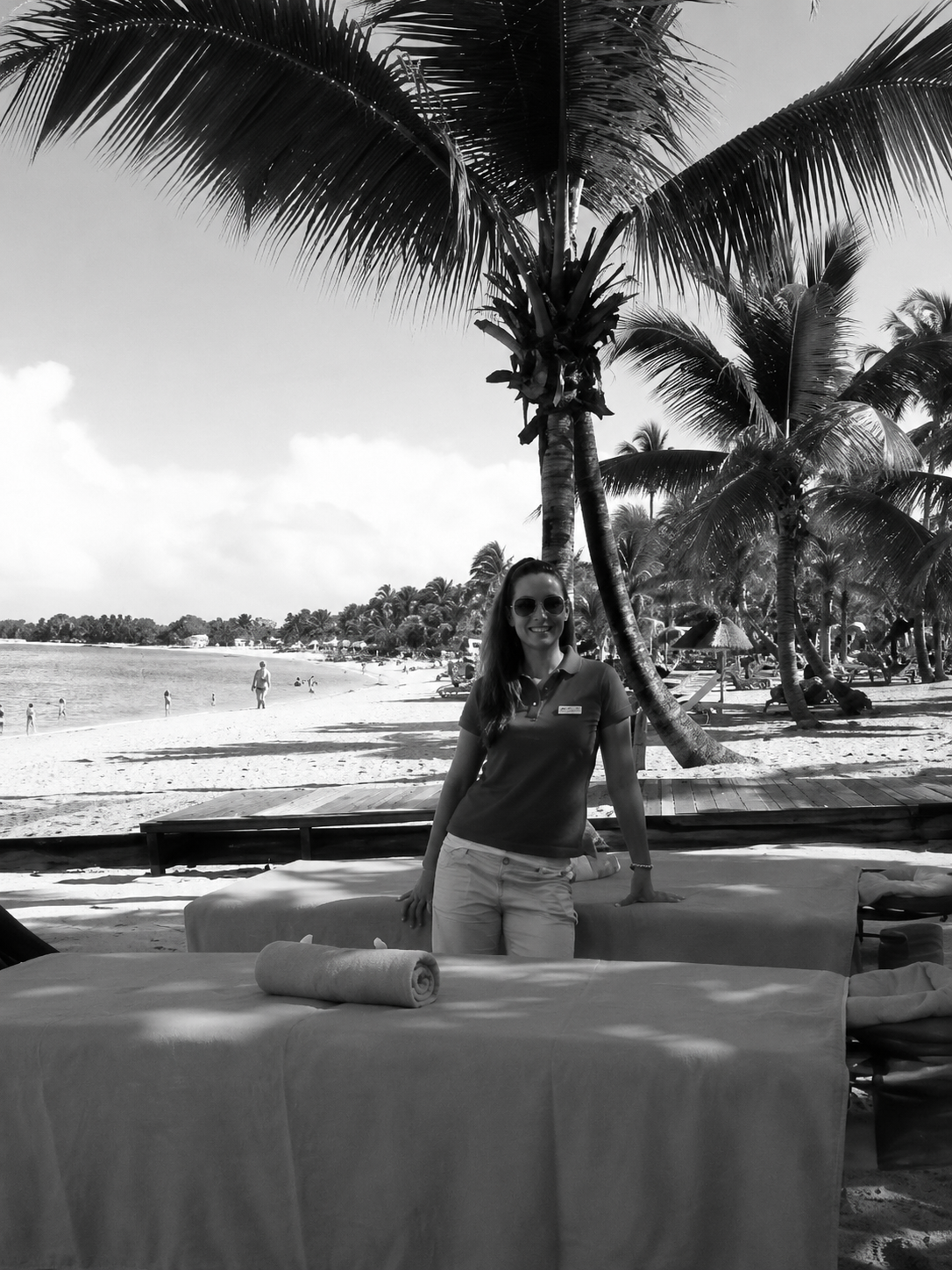 Die Kosmetikerin Johanna Arnemann steht in einem Polohemd und mit Sonnenbrille hinter zwei Massagetischen an einem Strand unter Palmen.
