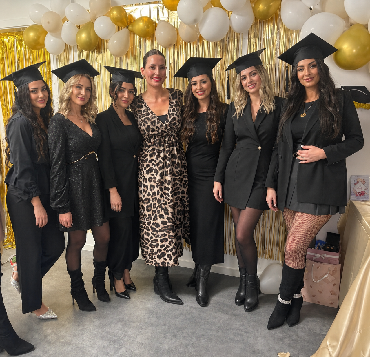 Sieben Absolventinnen der Kosmetikausbildung bei Johanna Arnemann posieren mit ihren Abschlusskappen vor einem Hintergrund mit goldenen Fransen für ein Foto.